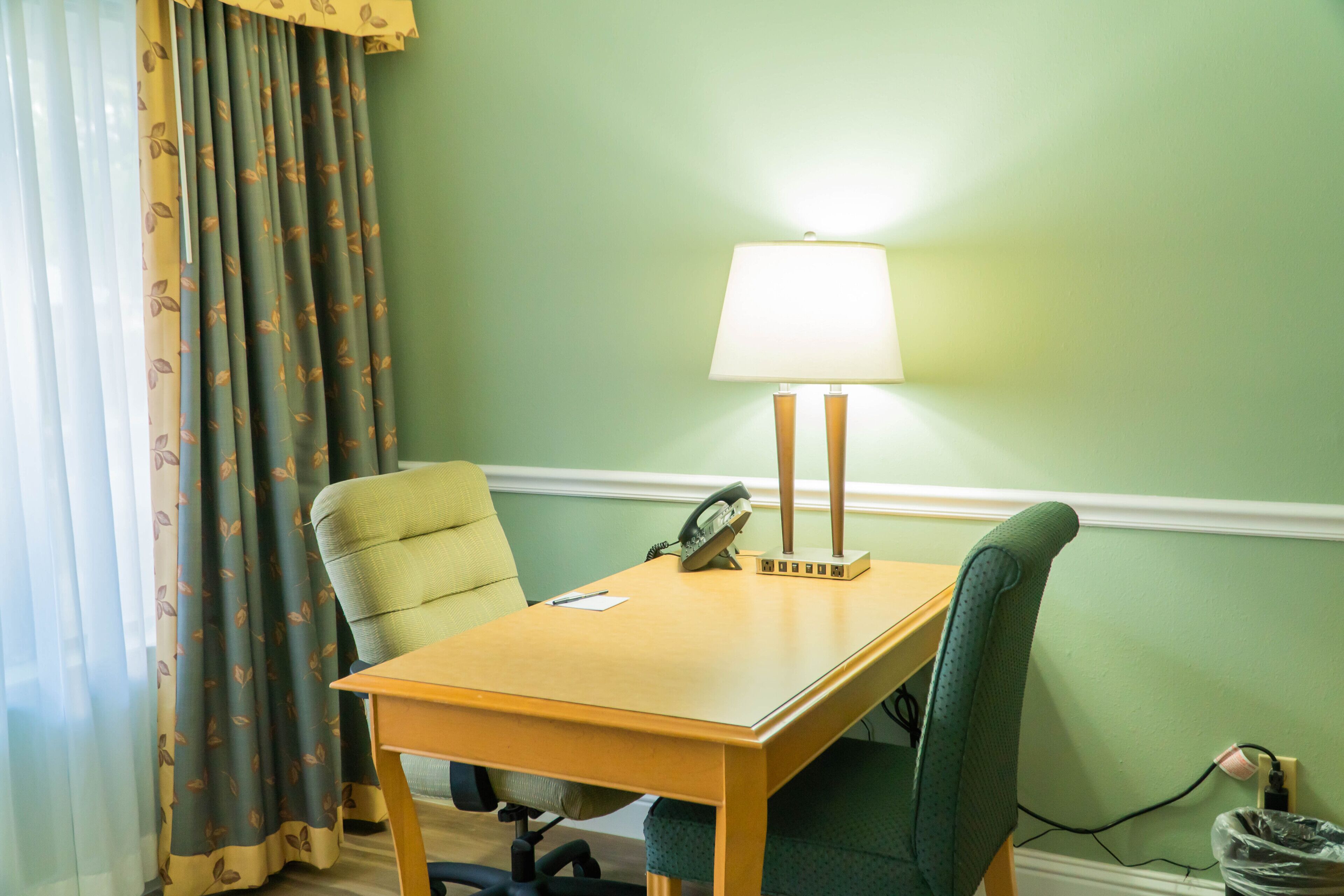 A hotel room with a wooden desk, two chairs, a lamp, a telephone, and a power outlet.