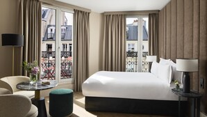 Habitación de hotel con una cama grande, una mesa redonda, una silla, un pequeño otoman y vistas de la ciudad a través de ventanas amplias.
