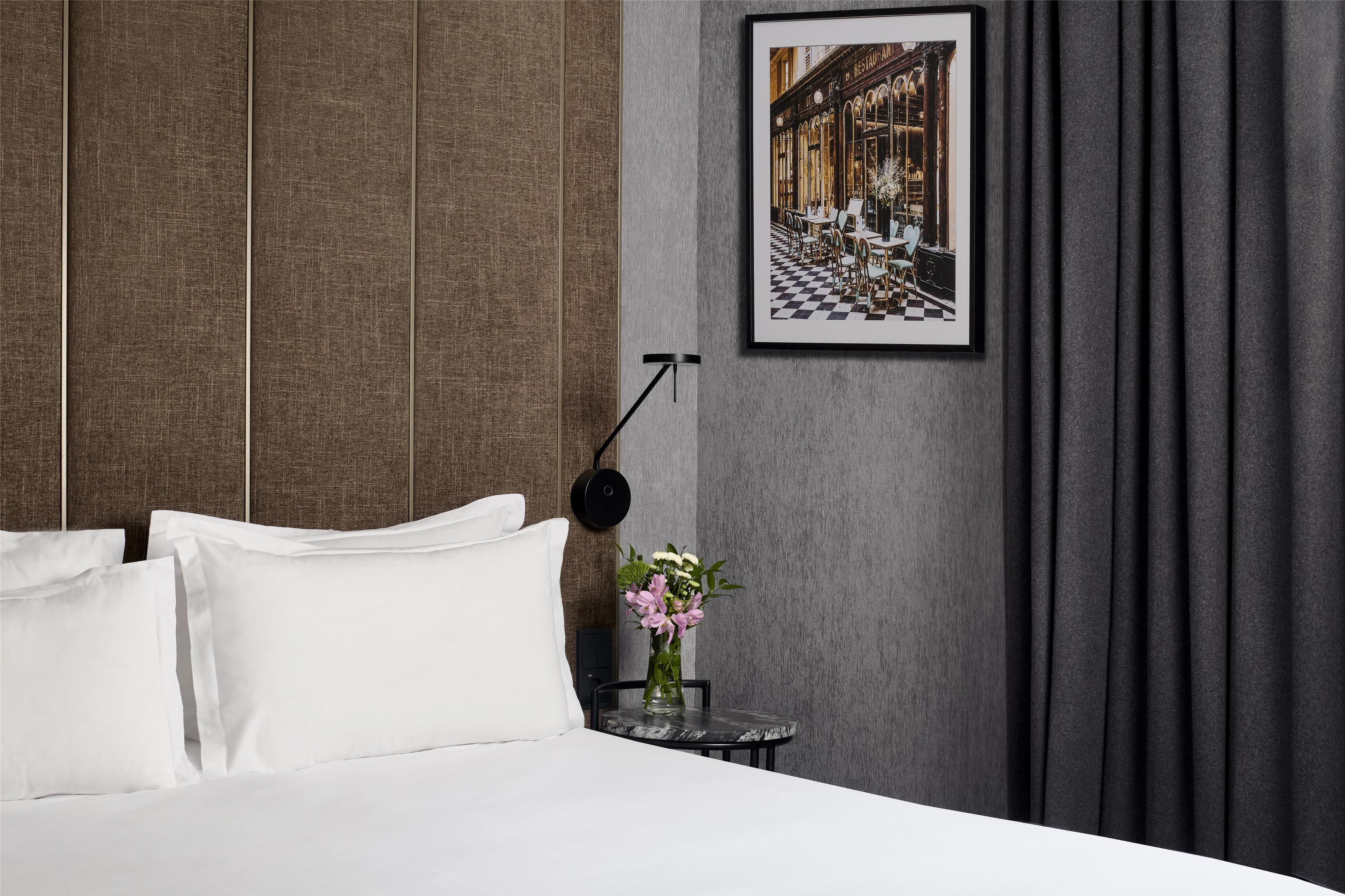A hotel room with a bed, white pillows, a small round table with a vase of flowers, a framed picture on the wall, and a black floor lamp.