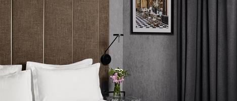 A hotel room with a bed, white pillows, a small round table with a vase of flowers, a framed picture on the wall, and a black floor lamp.