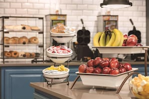 A buffet setup with various food items including donuts, pastries, fruits, and a bowl of fruit salad.