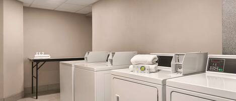 A laundry room with three white washers and dryers, a small table with a towel dispenser, and a grey textured wall.