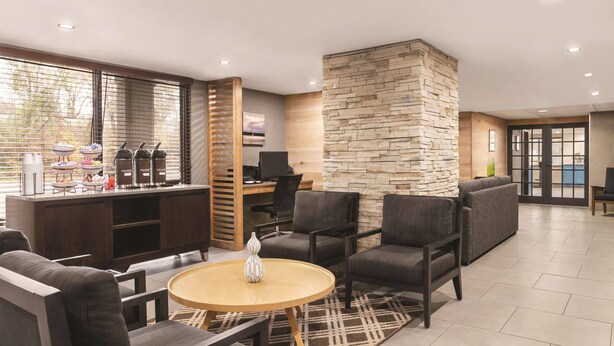 A modern hotel lobby with a stone accent wall, comfortable seating, a small round table, and a kitchenette area. - Country Inn & Suites by Radisson, Williamsburg East (Busch Gardens), VA (Williamsburg)