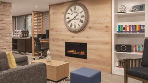 A modern living room with a fireplace, a large wall clock, and a bookshelf. - Country Inn & Suites by Radisson, Williamsburg East (Busch Gardens), VA (Williamsburg)