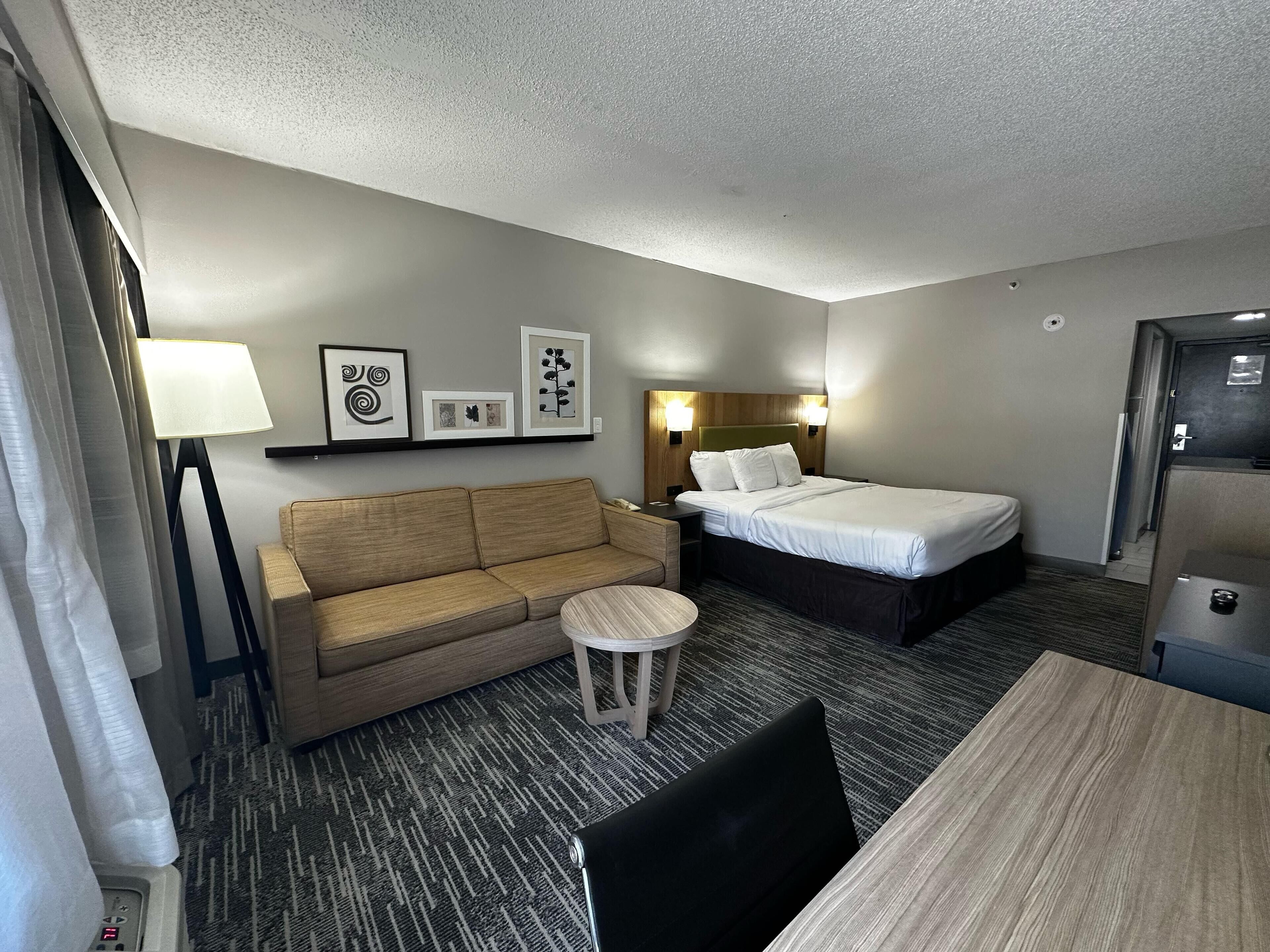 A hotel room with a bed, a sofa, a small round table, a lamp, and framed pictures on the wall.