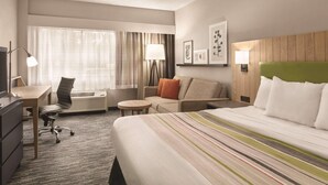 A hotel room with a large bed, a desk, a chair, a sofa, and a small table. There is a window with curtains, a lamp, and framed pictures on the wall. - Country Inn & Suites by Radisson, Williamsburg East (Busch Gardens), VA (Williamsburg)