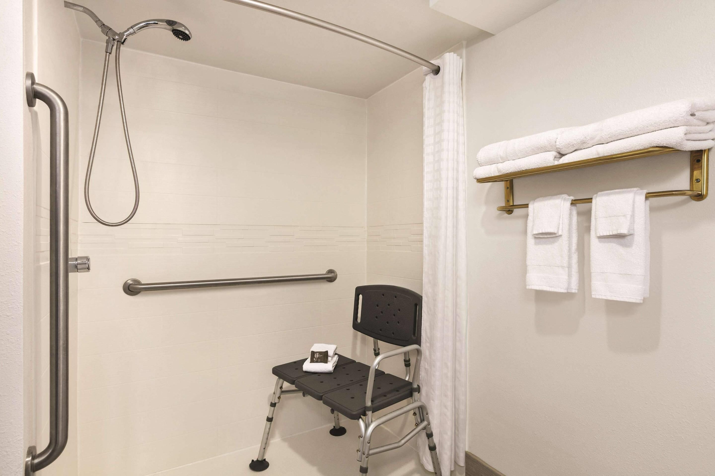 Un baño con ducha, barras de apoyo, un asiento accesible para silla de ruedas y un perchero para toallas.
