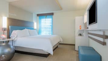 A hotel room with a bed, a desk with a lamp, a TV, and a small seating area.