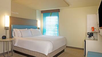A hotel room with a bed, bedside lamps, a nightstand, a window with curtains, and a small desk with a kettle and cups.