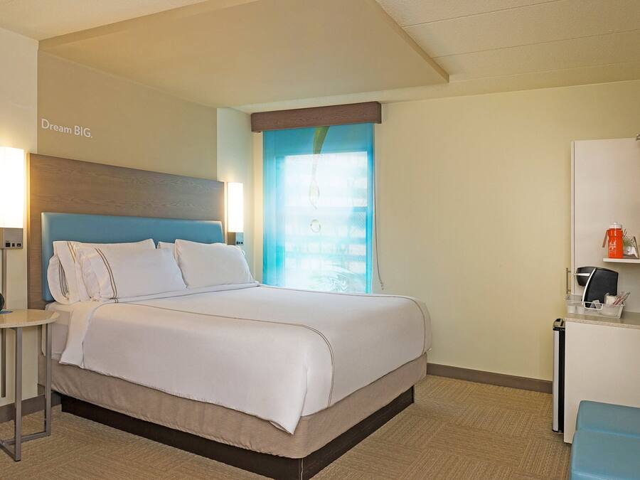 A hotel room with a bed, bedside lamps, a nightstand, a window with curtains, and a small desk with a kettle and cups.