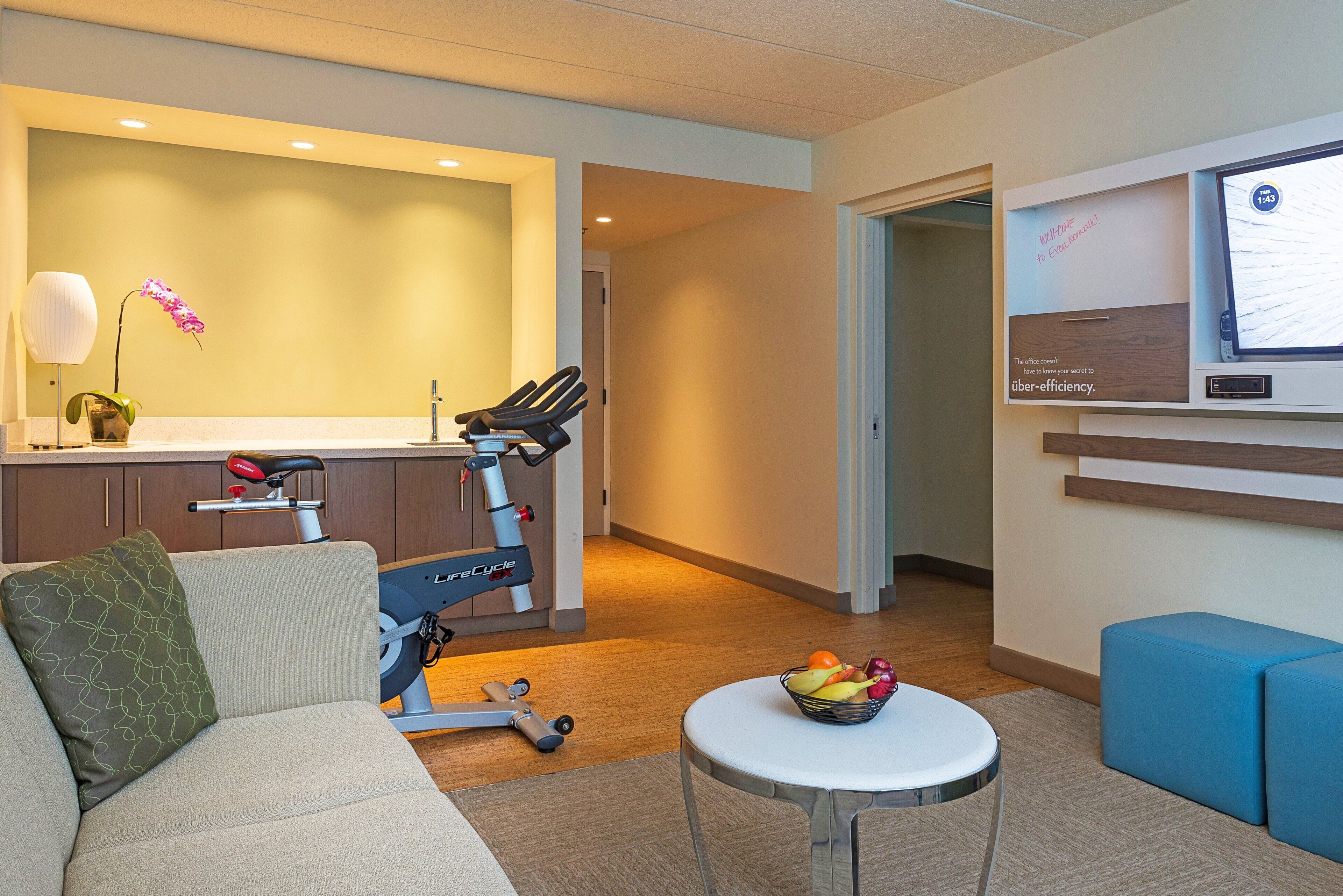 A modern office space with a treadmill, a desk with a computer, a couch, and a small table with fruit.
