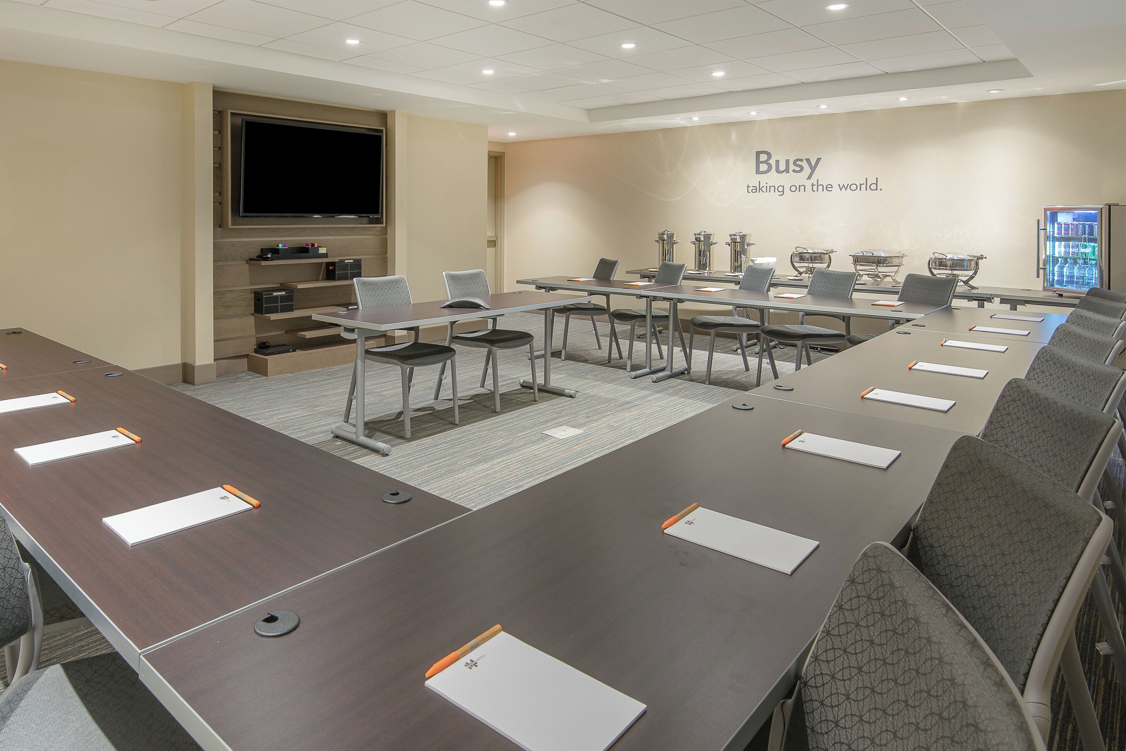A conference room with a long table, chairs, a television, and a wall with the text 'Busy taking on the world.'