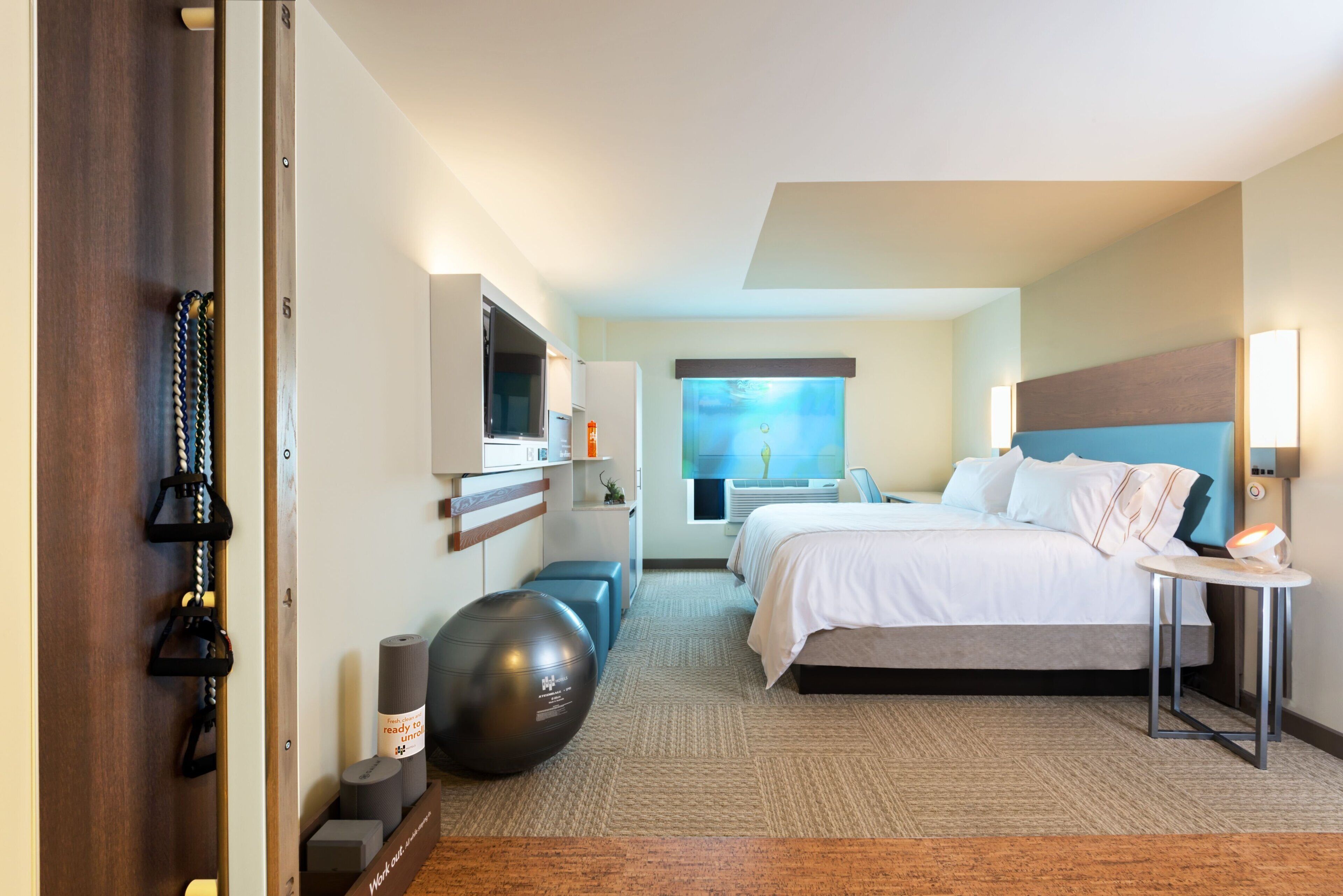 A hotel room with a bed, a desk, a chair, a TV, a workout ball, and a window with a view.