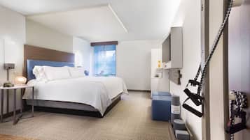 Habitación de hotel con cama, mesita de noche, lámpara, televisor, equipo de ejercicio y ventana con vistas.