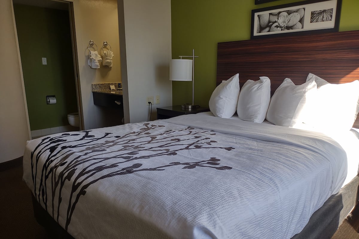 Habitación de hotel con una cama grande, una mesita de noche, una lámpara de pared y un baño visible a través de una puerta abierta.