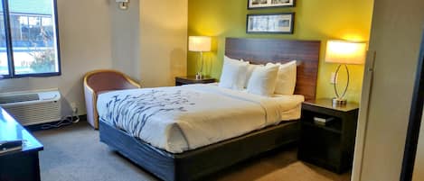 A hotel room with a large bed, two bedside tables with lamps, a chair, and a window with an air conditioner.