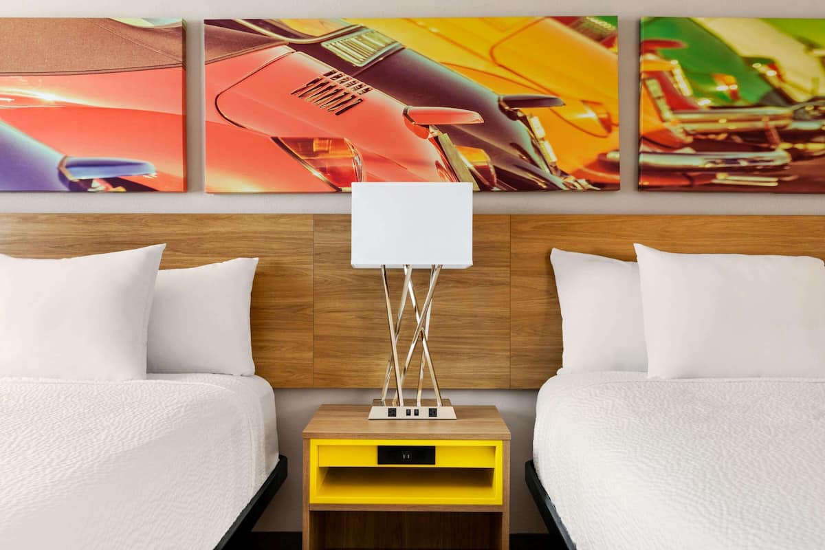 A hotel room with two beds, a wooden headboard, a nightstand with a lamp, and a wall with three colorful paintings.