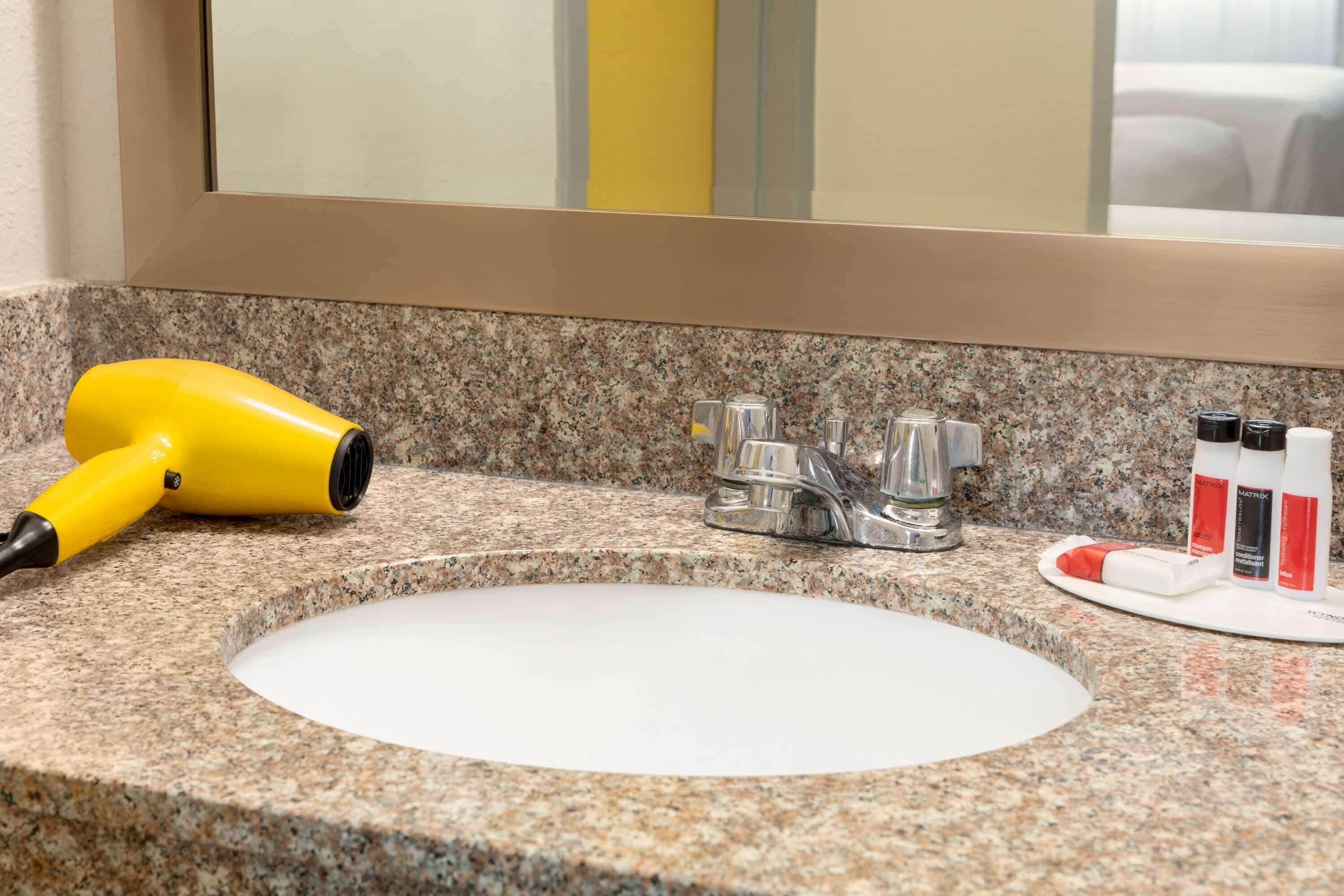 A bathroom with a granite countertop, a white sink, a yellow hair dryer, and toiletries on the counter.
