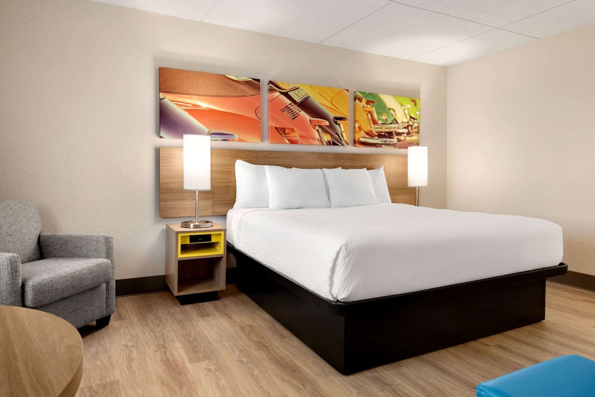A modern hotel room with a large bed, a gray armchair, a small table, and a wall-mounted artwork.