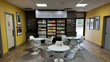 Una zona de estar con mesas y sillas blancas, un refrigerador lleno de bebidas y cuadros en las paredes.
