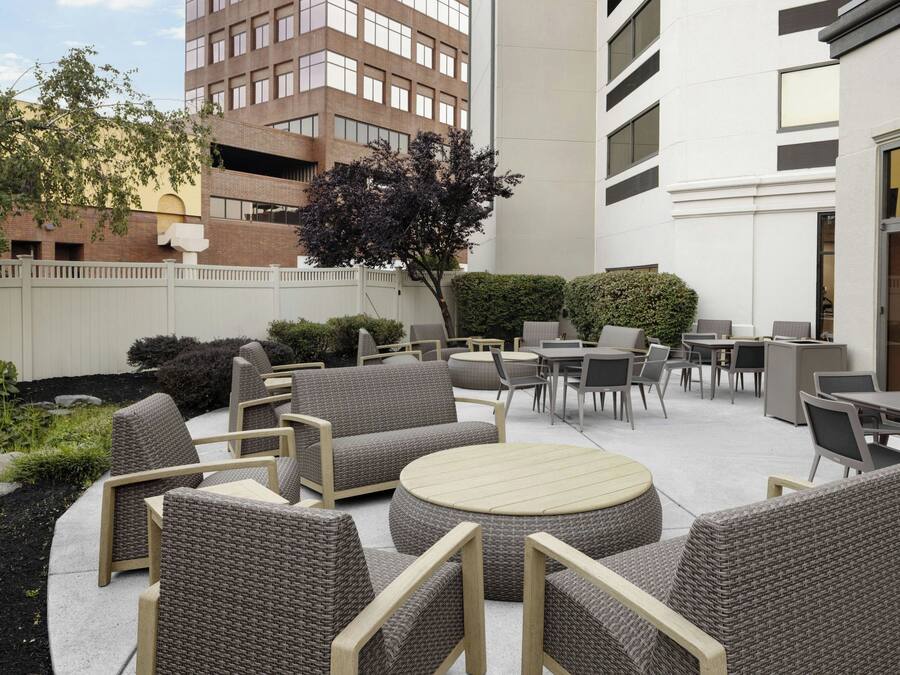 Een terras buiten met rieten meubels, waaronder banken en tafels, omgeven door een lage witte hek en begroeiing.
