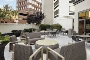 Ein Terrassenbereich im Freien mit Korbmöbeln, darunter Sofas und Tische, umgeben von einem niedrigen weißen Zaun und viel Grün.