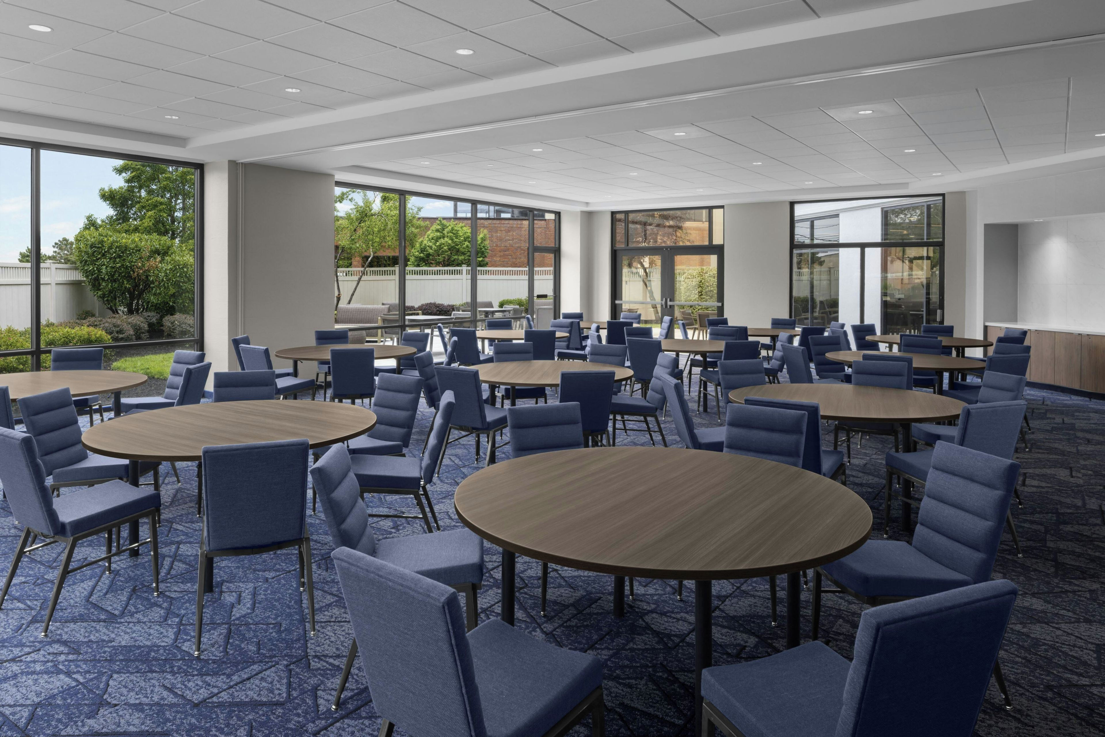 A conference room with round tables and chairs, large windows, and a carpeted floor.