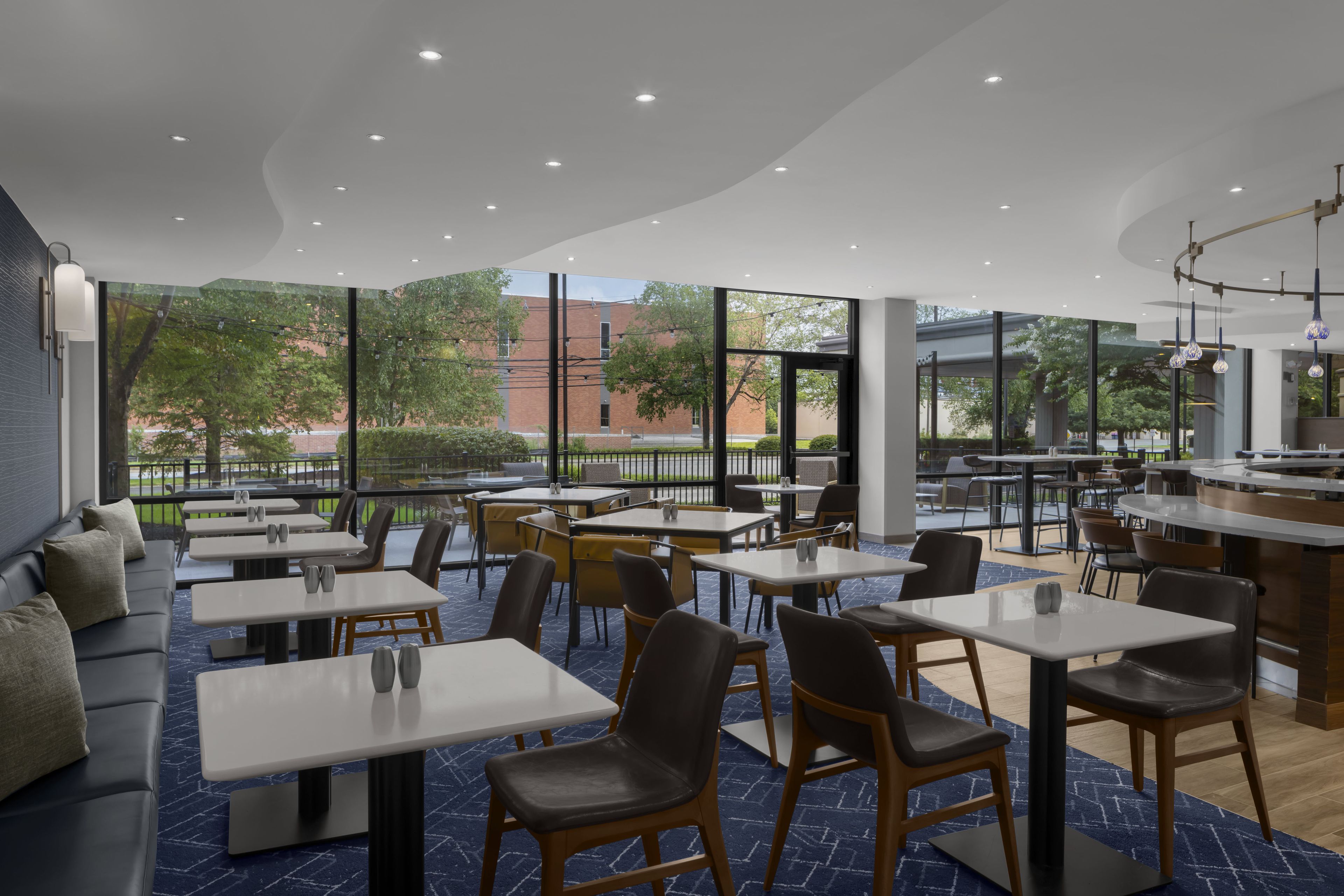 Un restaurante moderno con distintas opciones de asientos, incluyendo mesas y sofás, con amplios ventanales que ofrecen vistas al exterior.