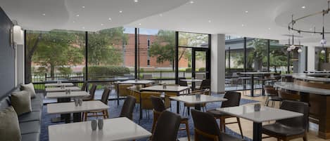 A modern restaurant with a mix of seating options, including tables and a sofa, with large windows offering a view of the outdoors.
