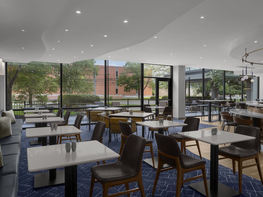 Ein modernes Restaurant mit verschiedenen Sitzgelegenheiten, darunter Tische und eine Sitzecke, große Fenster mit Blick ins Freie.