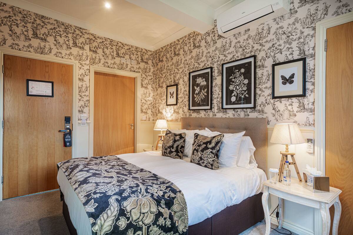 Een hotelkamer met een groot bed, een nachtkastje met een lamp, een ingelijst kunstwerk aan de muur en een deur met een bordje.