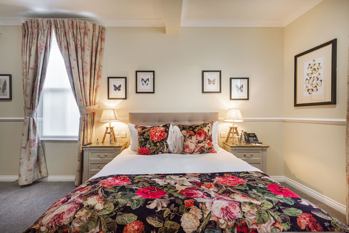 Een slaapkamer met een bloemenbedekker, hoofdbord, nachtlampjes, een telefoon en vlinderkunst in een lijst aan de muur.