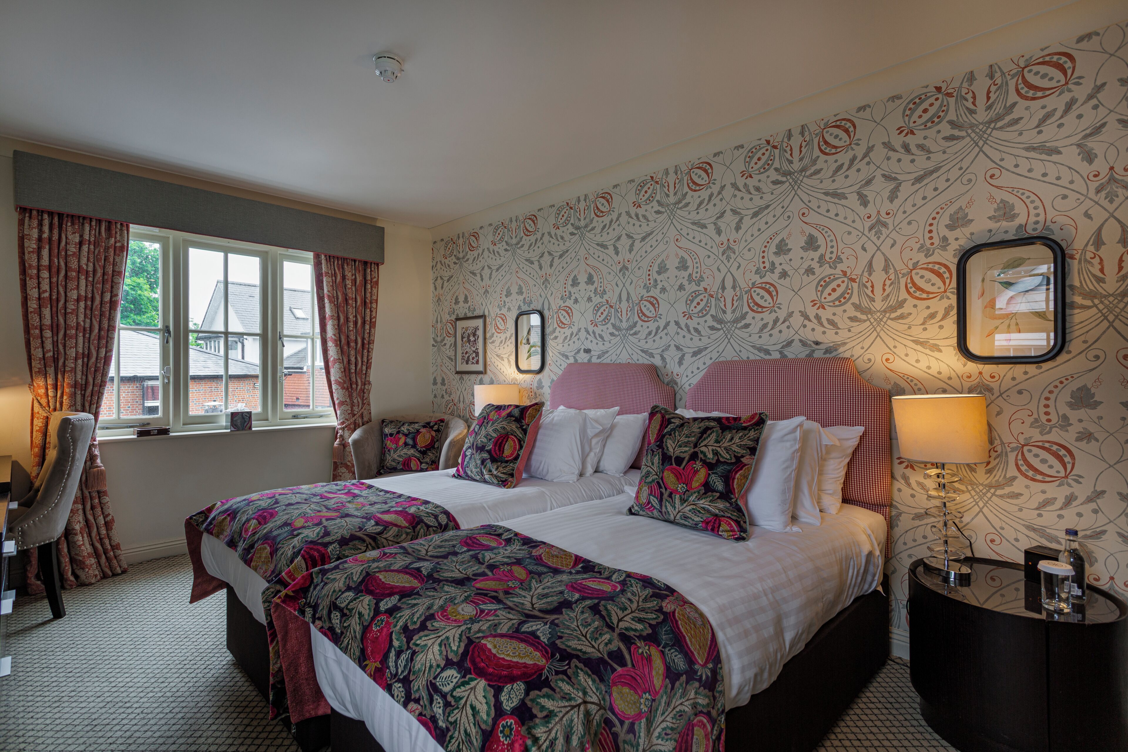 A double bed with floral bedding, a bedside table with a lamp, a chair, and a window with curtains.