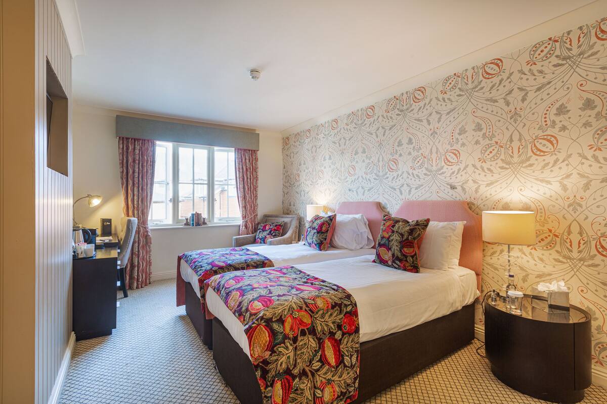 Een hotelkamer met twee bedden, een bureau, een stoel, een raam met gordijnen en behang met een patroon.