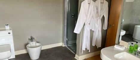 Un baño con ducha, lavamanos e inodoro. Hay dos albornoces blancos colgados en un perchero.