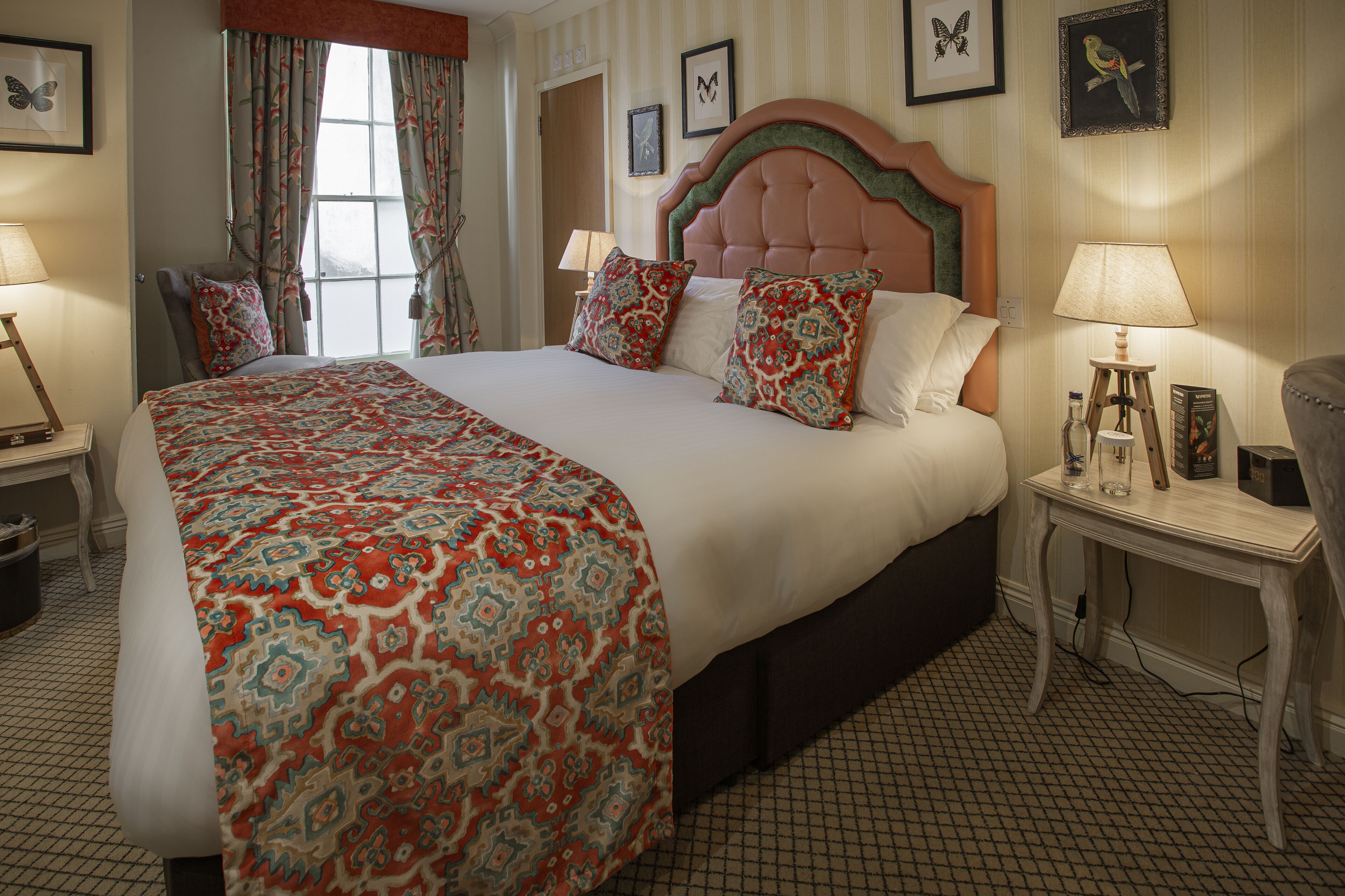 A large bed with a patterned bedspread, a wooden nightstand with a lamp, and a chair in a hotel room with striped wallpaper and framed pictures on the wall.