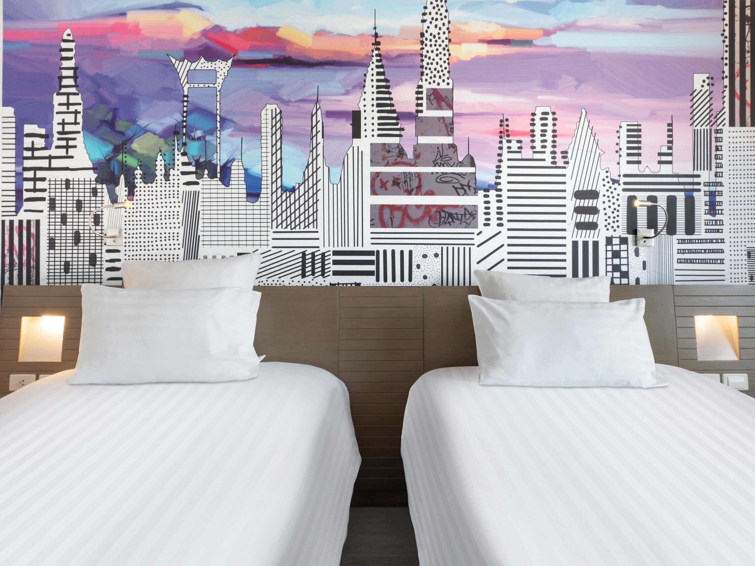 Habitación de hotel con dos camas, un mural de paisaje urbano en la pared y cabecera de madera.
