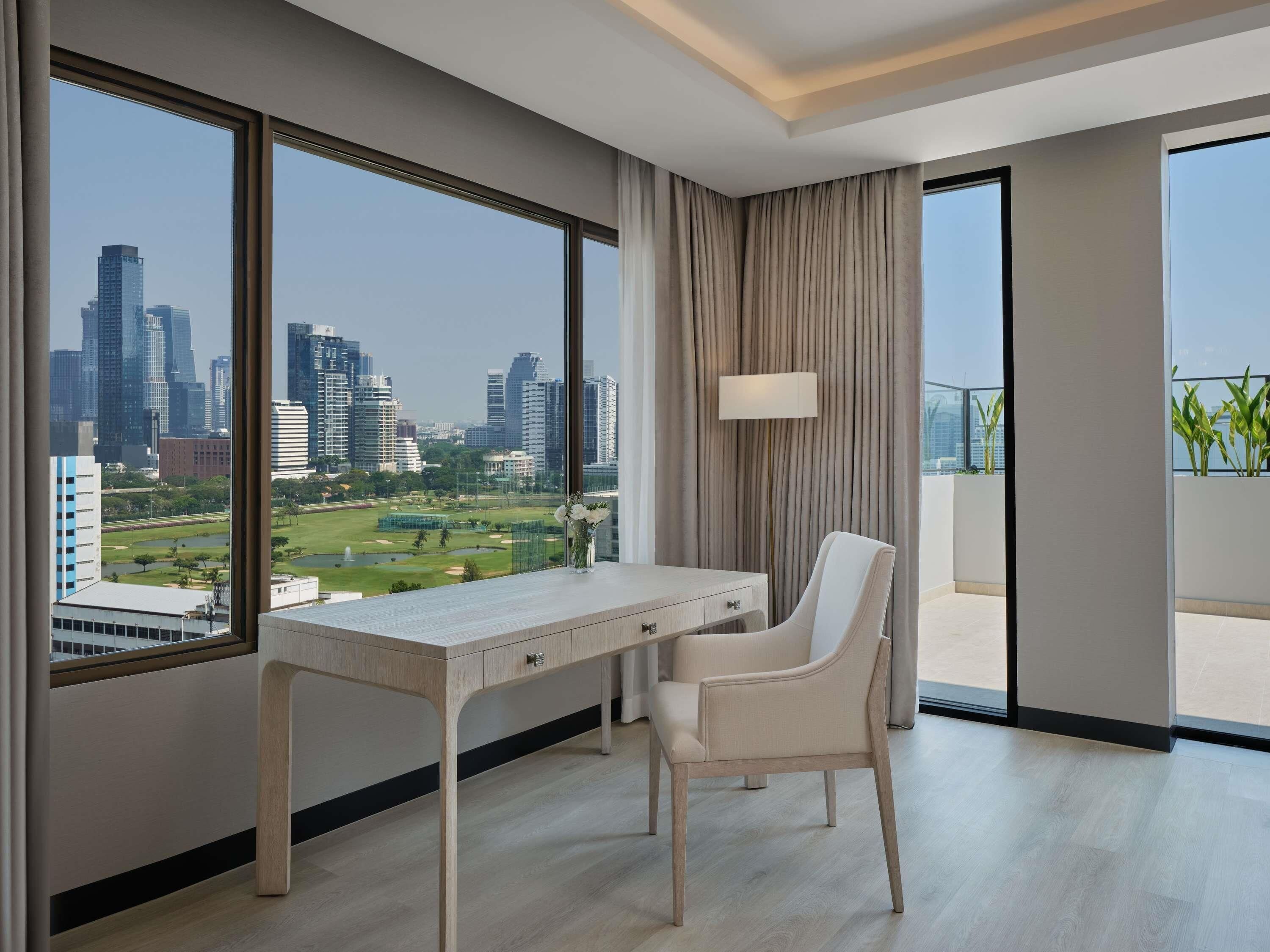 Une chambre d’hôtel moderne équipée d’un bureau, d’une chaise, de grandes fenêtres et offrant une vue sur le paysage urbain.