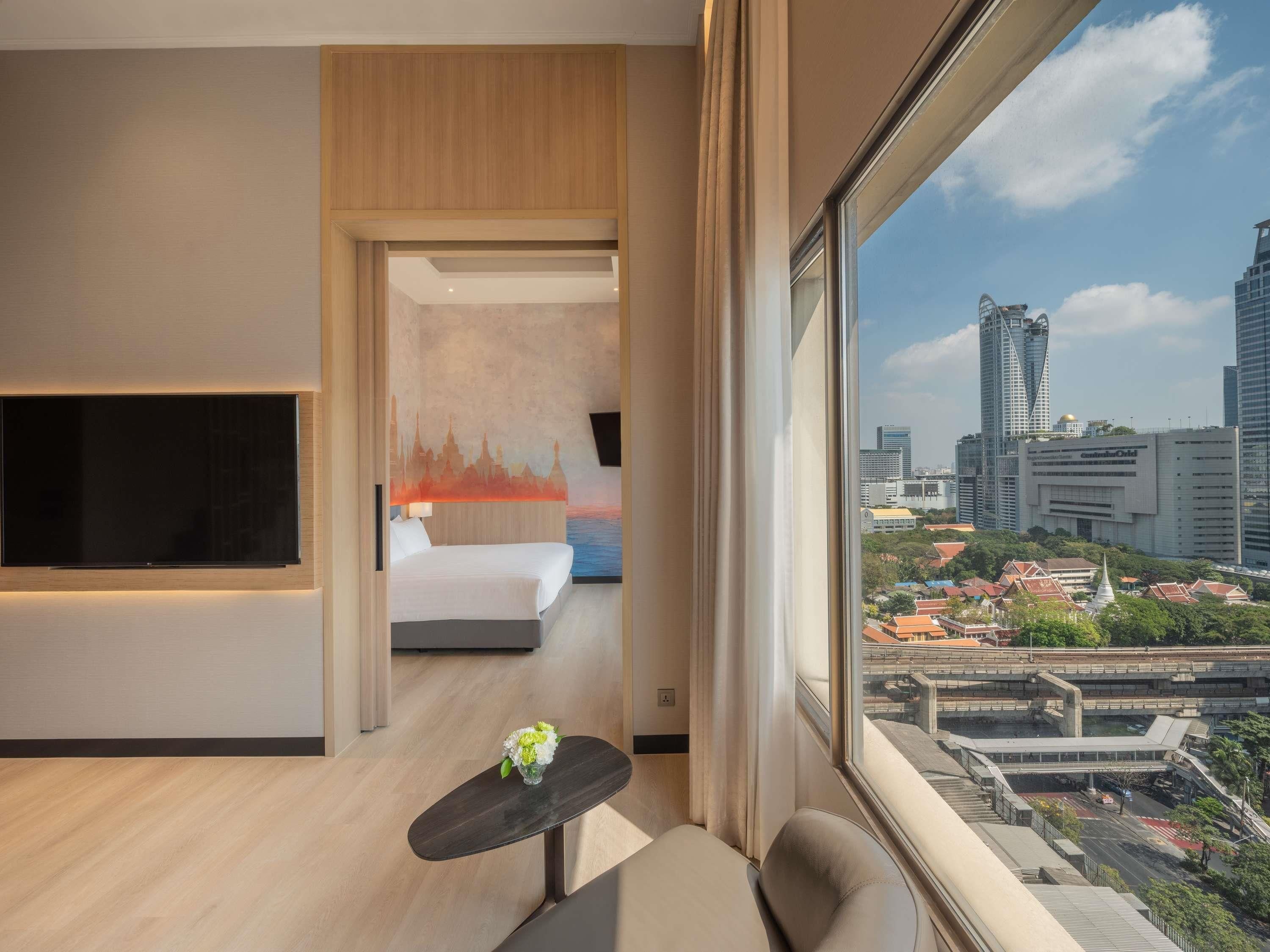 A modern hotel room with a large TV, a bed, a small table with a vase, and a city view.