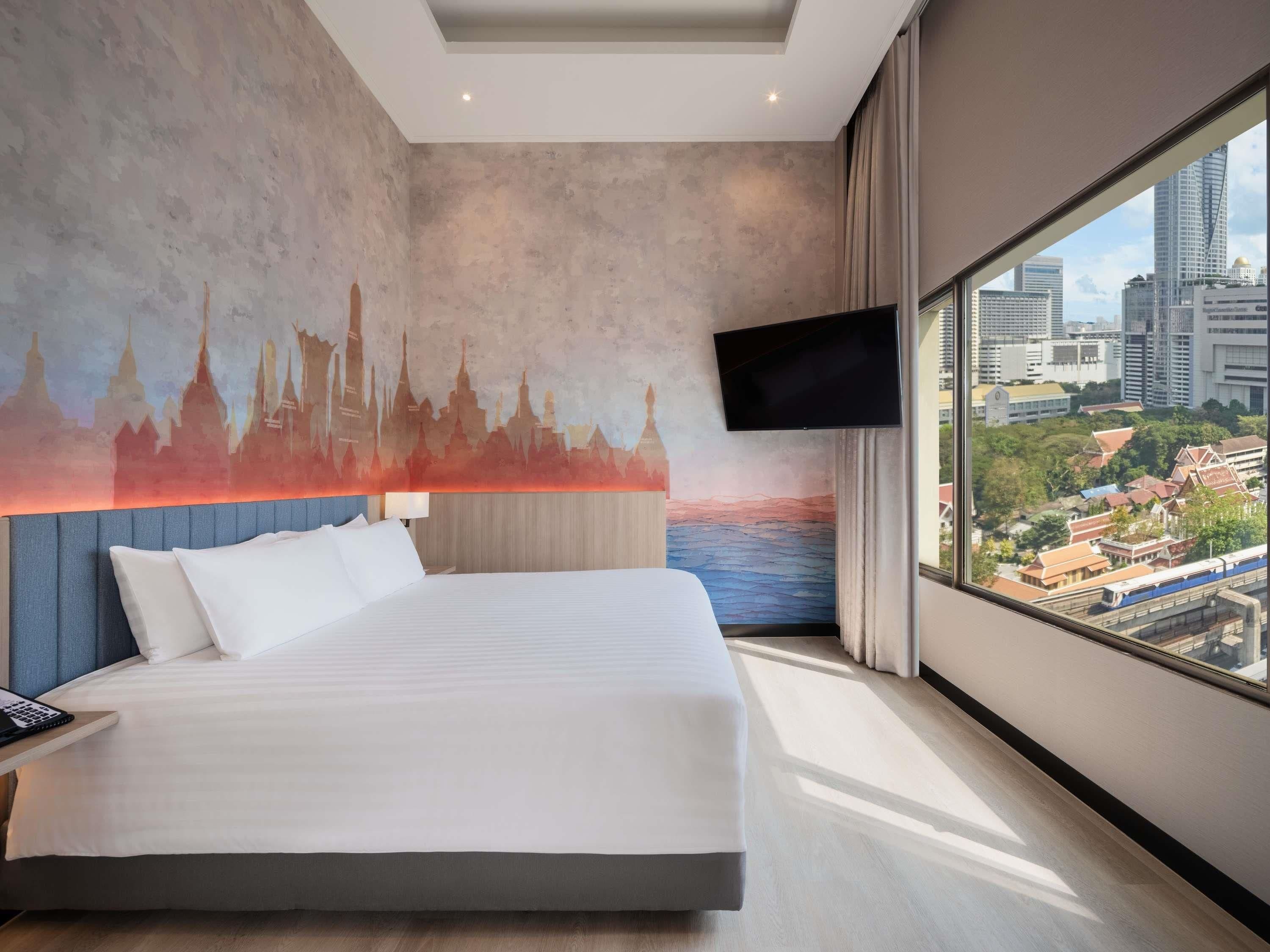 A modern hotel room with a large bed, a cityscape mural on the wall, a flat-screen TV, and a view of the city through large windows.