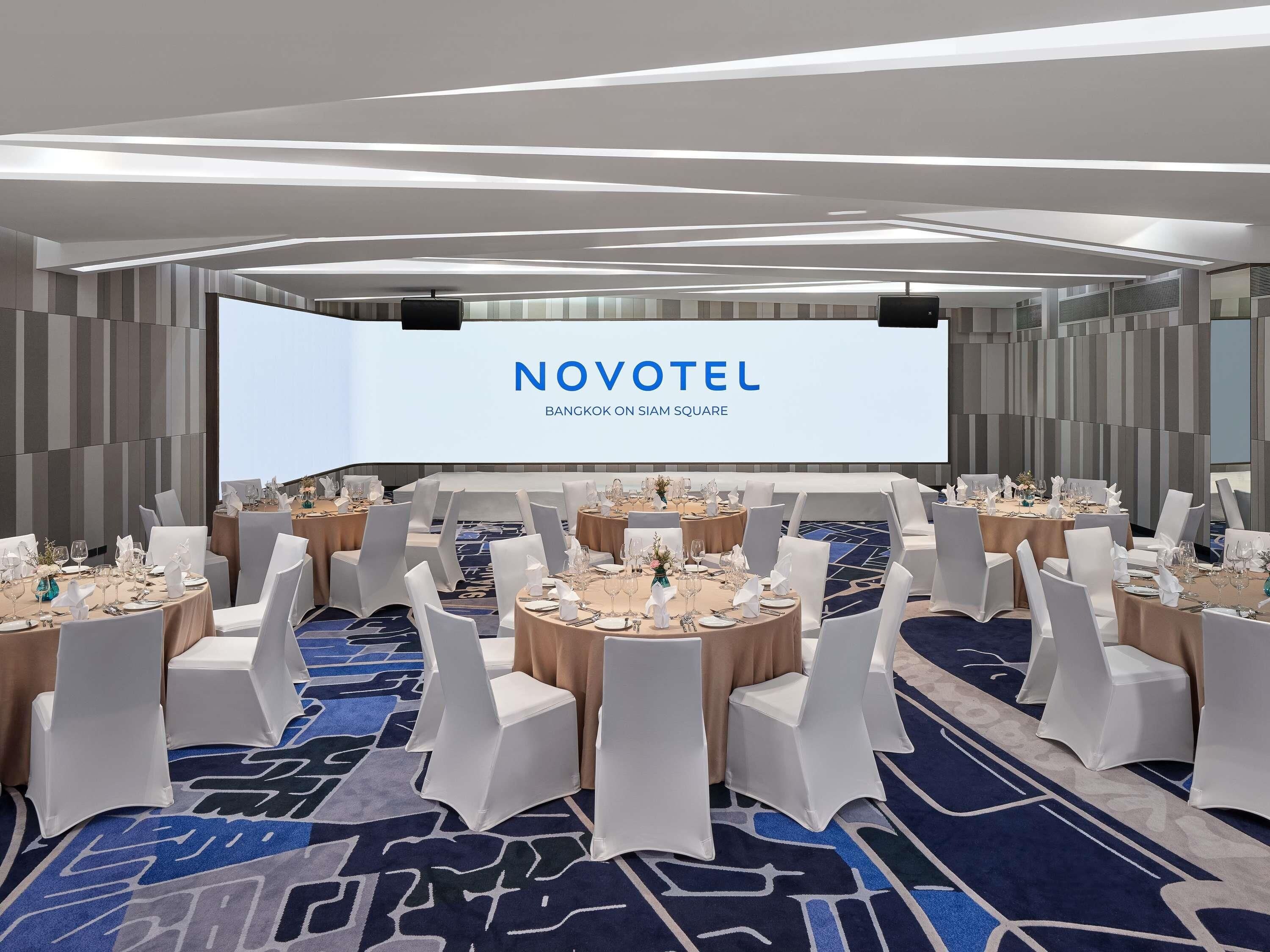 A Novotel hotel room with a large screen displaying the hotel's name and location, round tables with white chairs, and a patterned carpet.