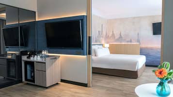 A modern hotel room with a bed, a desk with a flat-screen TV, a coffee machine, and a wall-mounted TV.