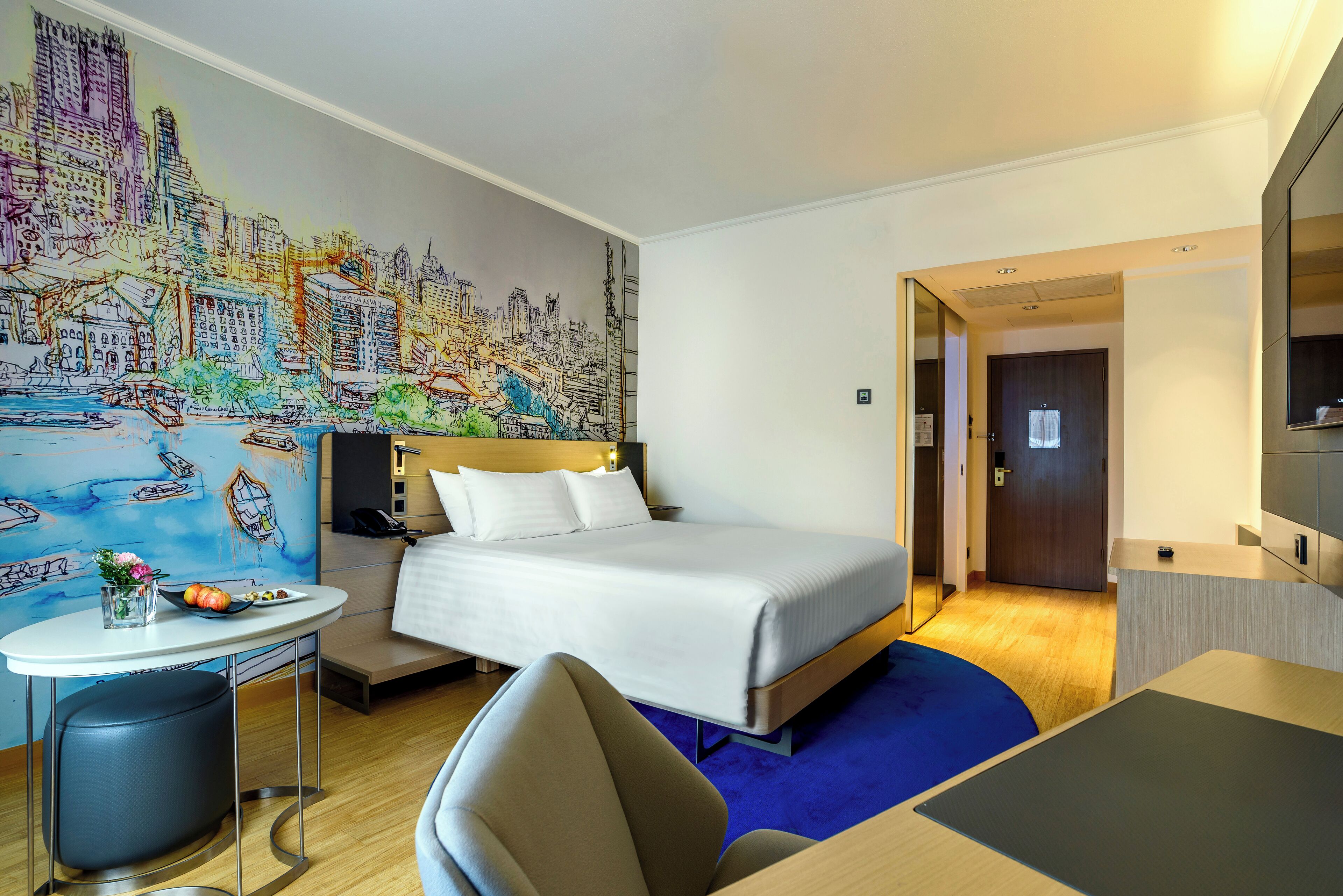 A hotel room with a bed, a desk, a chair, a small table with a vase and fruit, and a colorful cityscape mural on the wall.