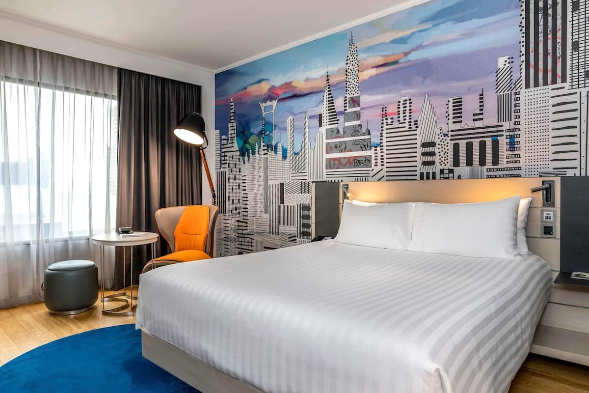 A hotel room with a bed, a chair, a lamp, a round table, and a cityscape mural on the wall.