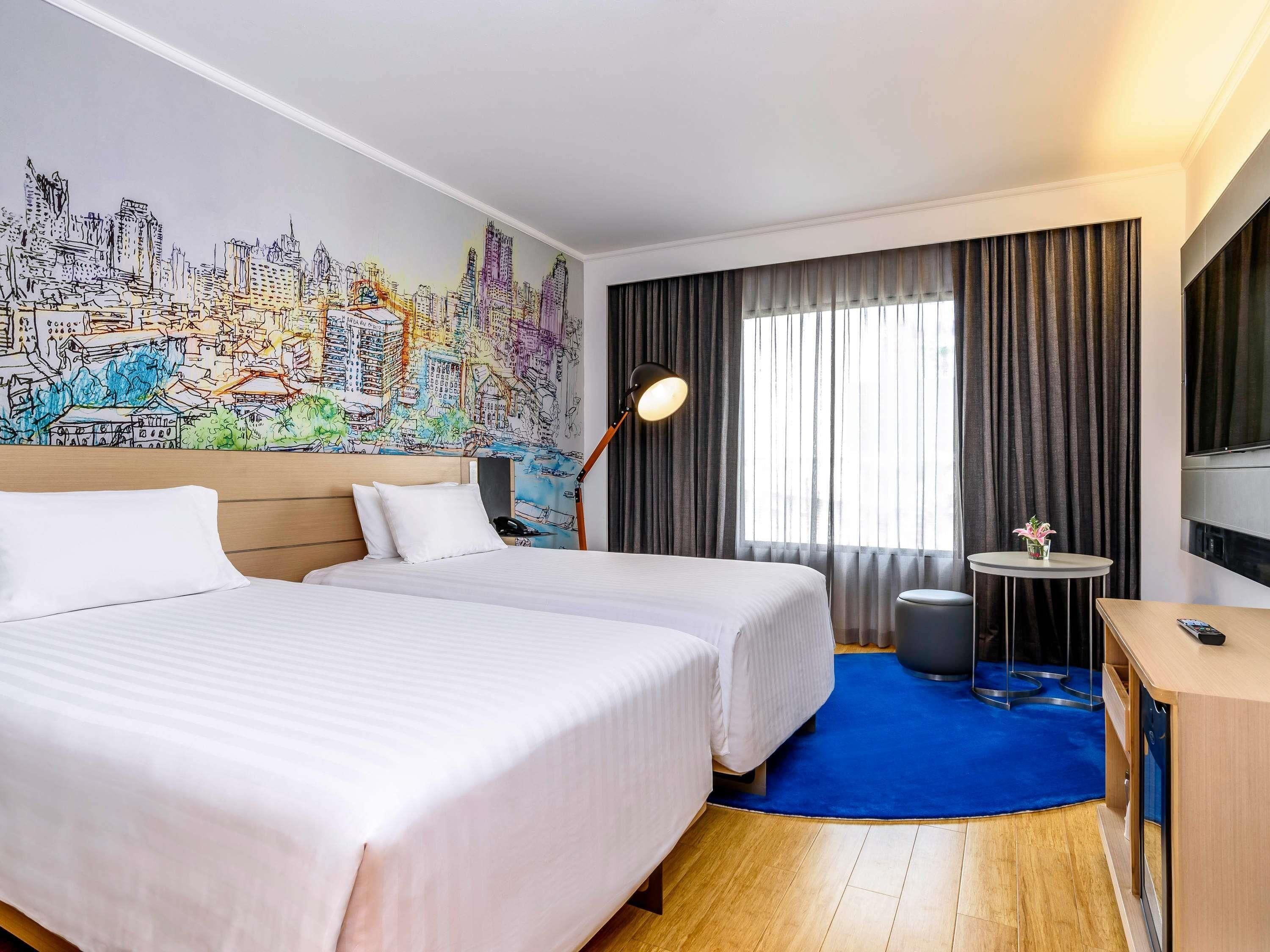 A hotel room with two beds, a cityscape mural, a desk with a chair, and a large window with curtains.