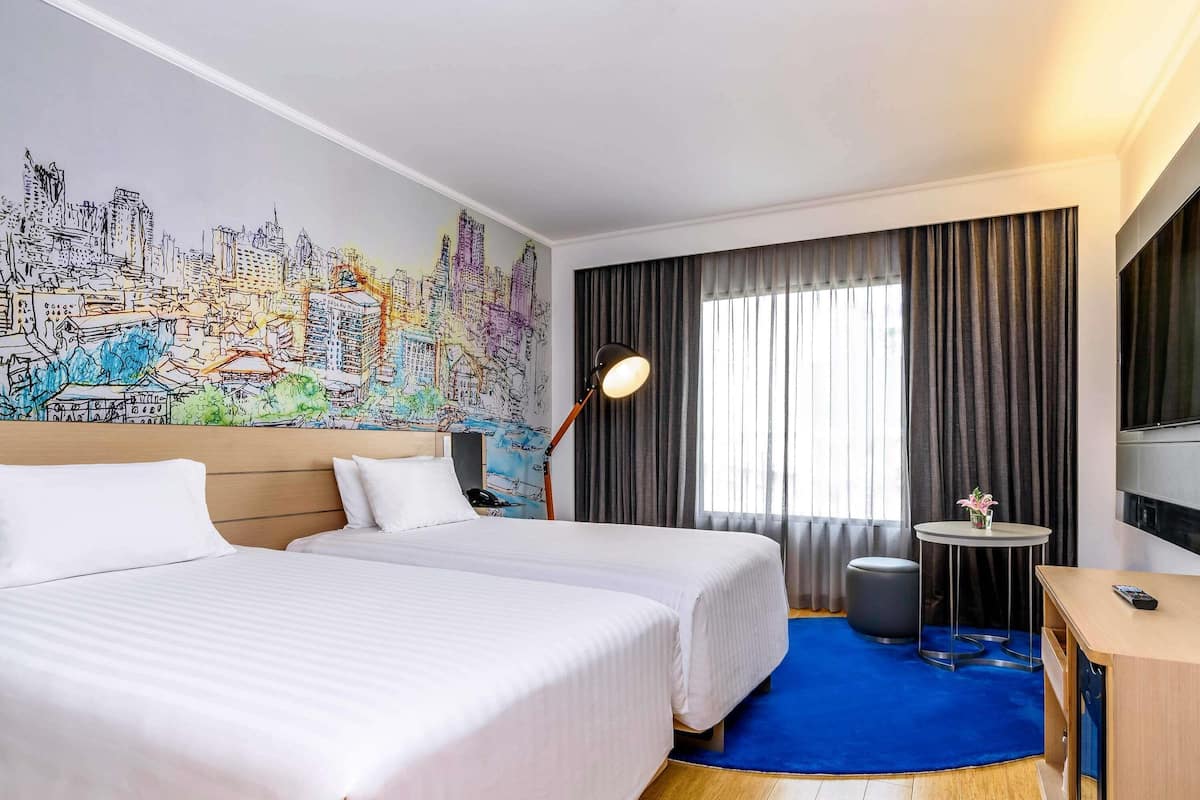 A hotel room with two beds, a cityscape mural, a desk with a chair, and a large window with curtains.