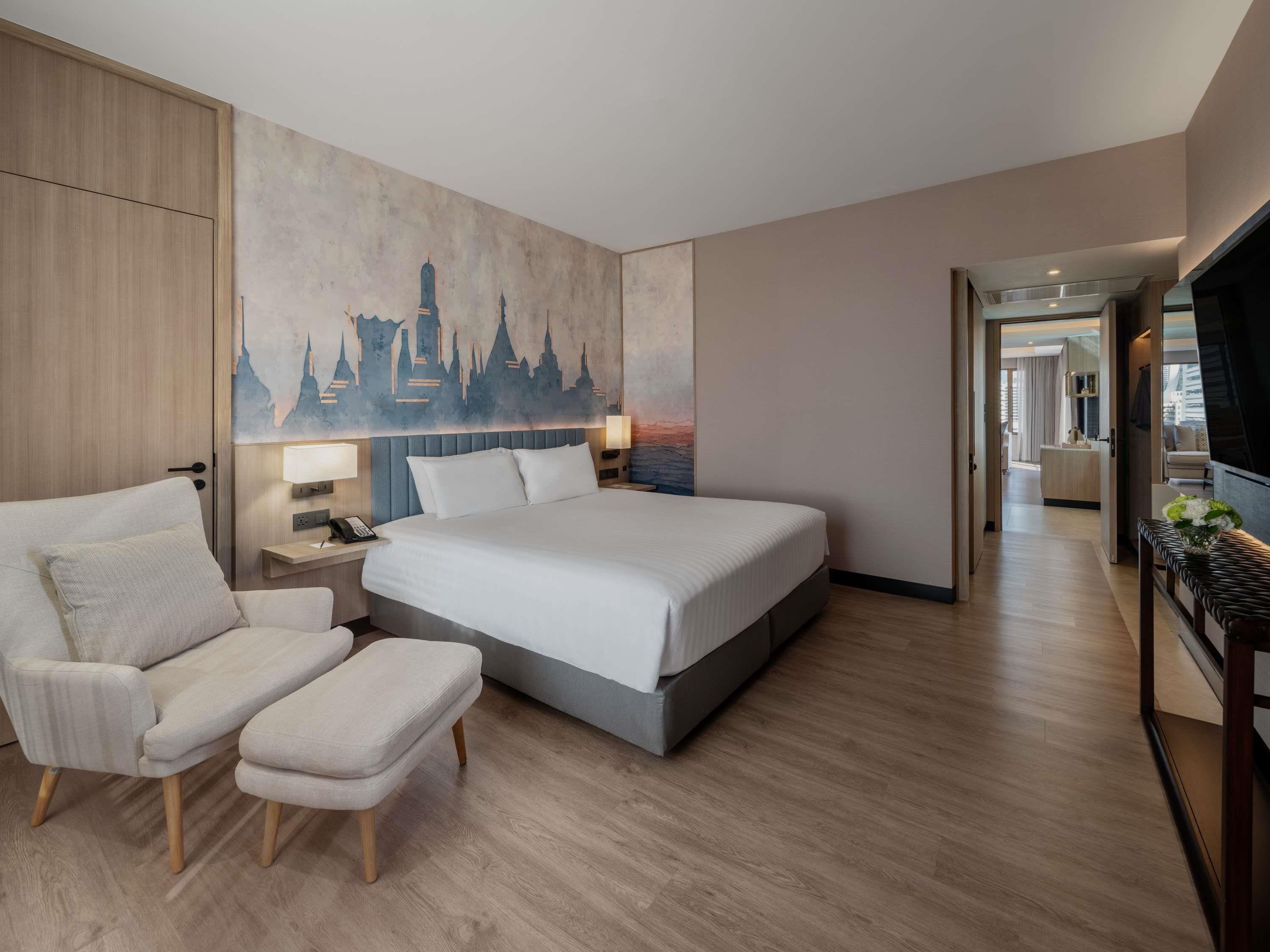 A modern hotel room with a large bed, a sitting area with a chair and ottoman, a nightstand with a lamp, and a painting of a city skyline on the wall.