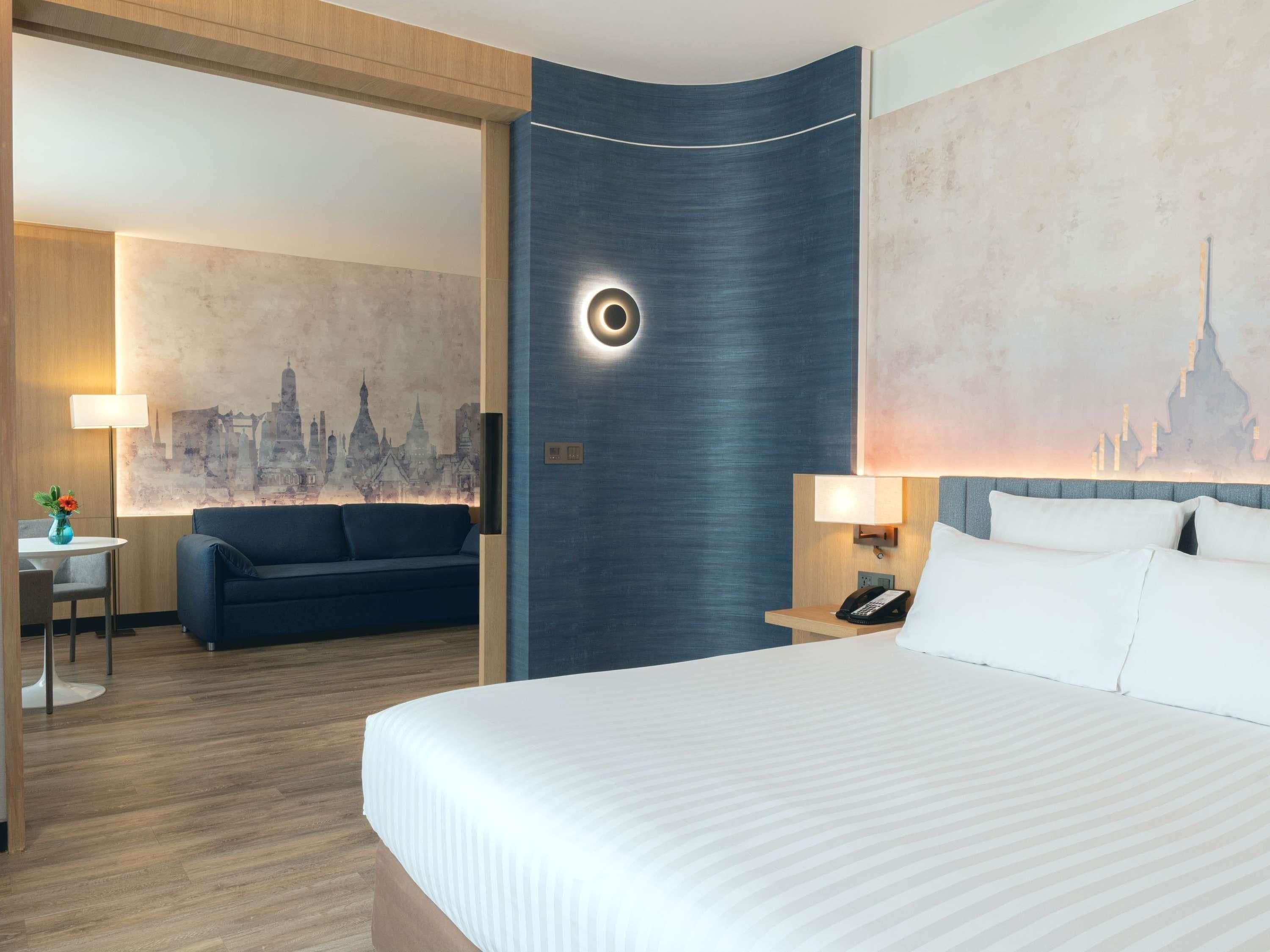 Habitación de hotel con cama, sofá, escritorio con lámpara y un mural de paisaje urbano en la pared.