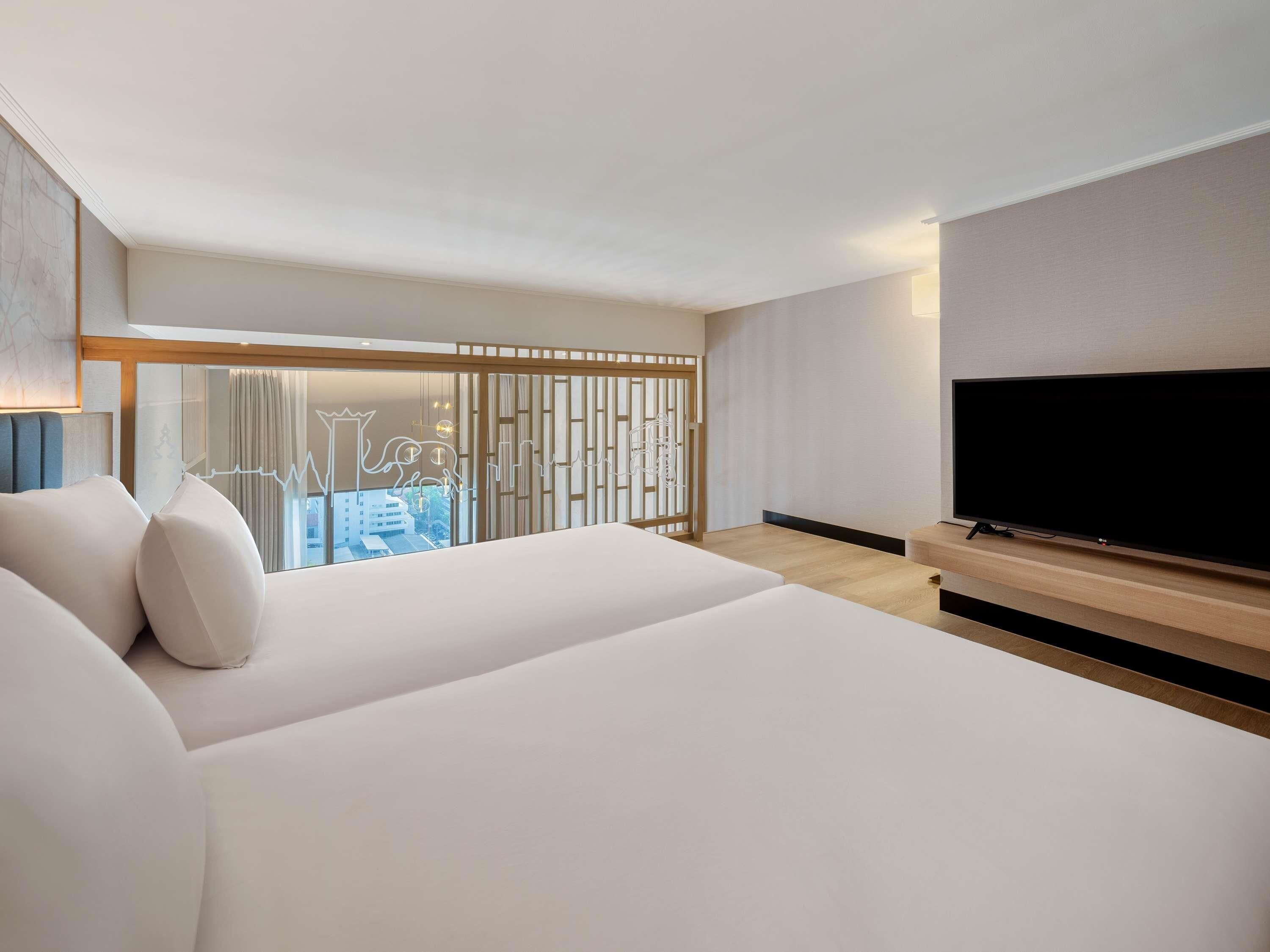 A modern hotel room with a large bed, a flat-screen TV, and a view of the city through a glass partition.
