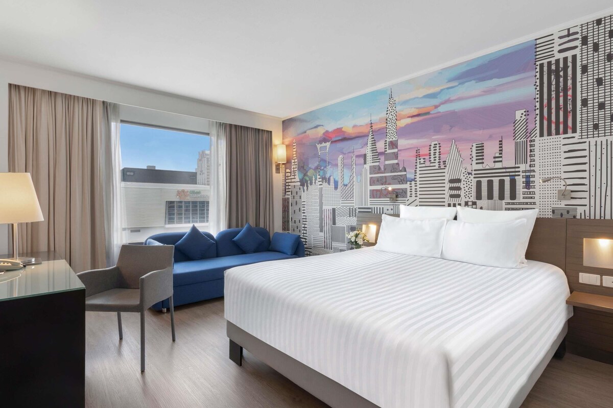 A hotel room with a large bed, a sofa, a desk, and a cityscape mural on the wall.